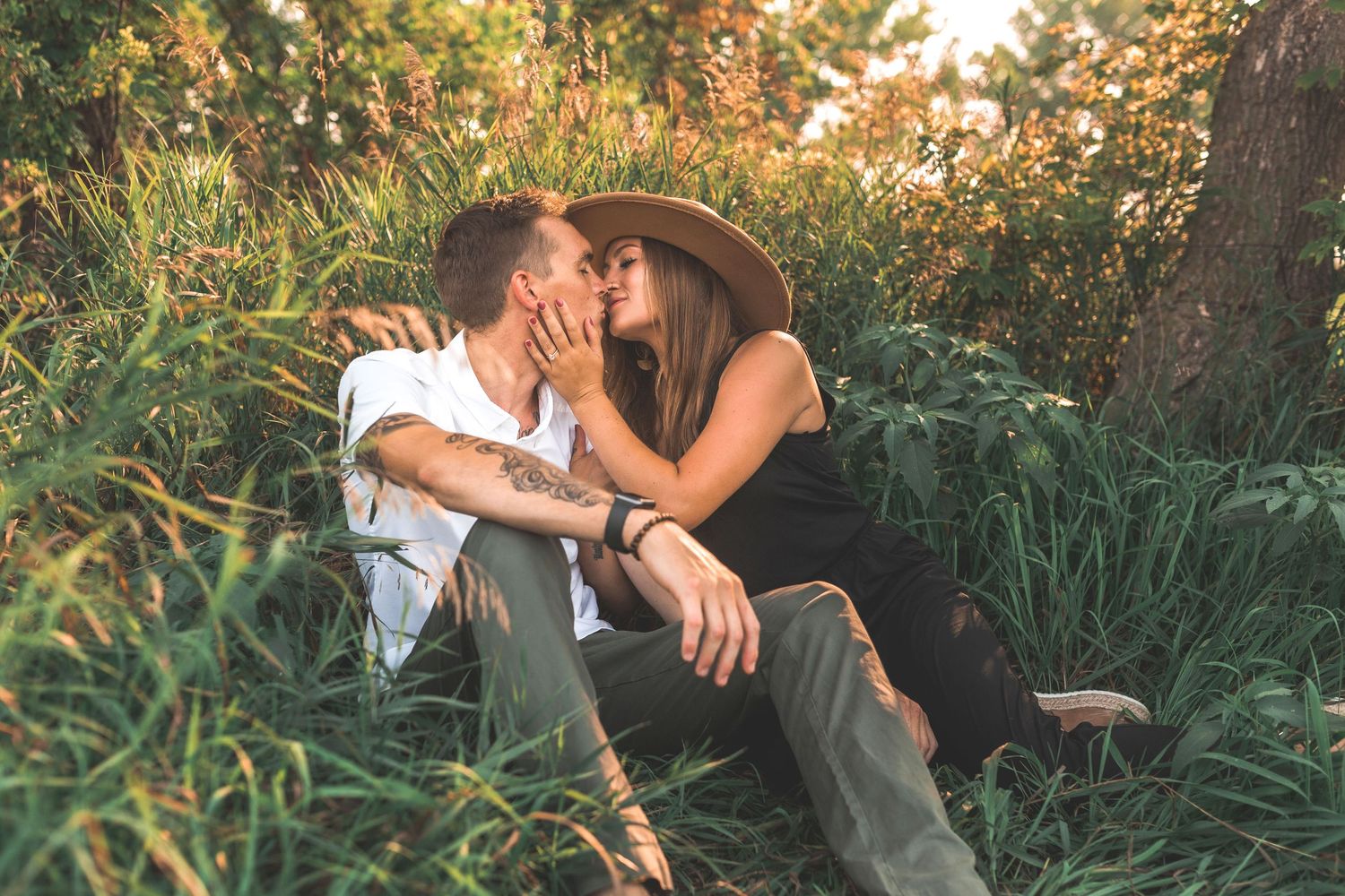 Infinity & Co. Photographer. A couple sharing a romantic kiss while sitting in lush green grass near a tree, woman wearing a stylish hat.