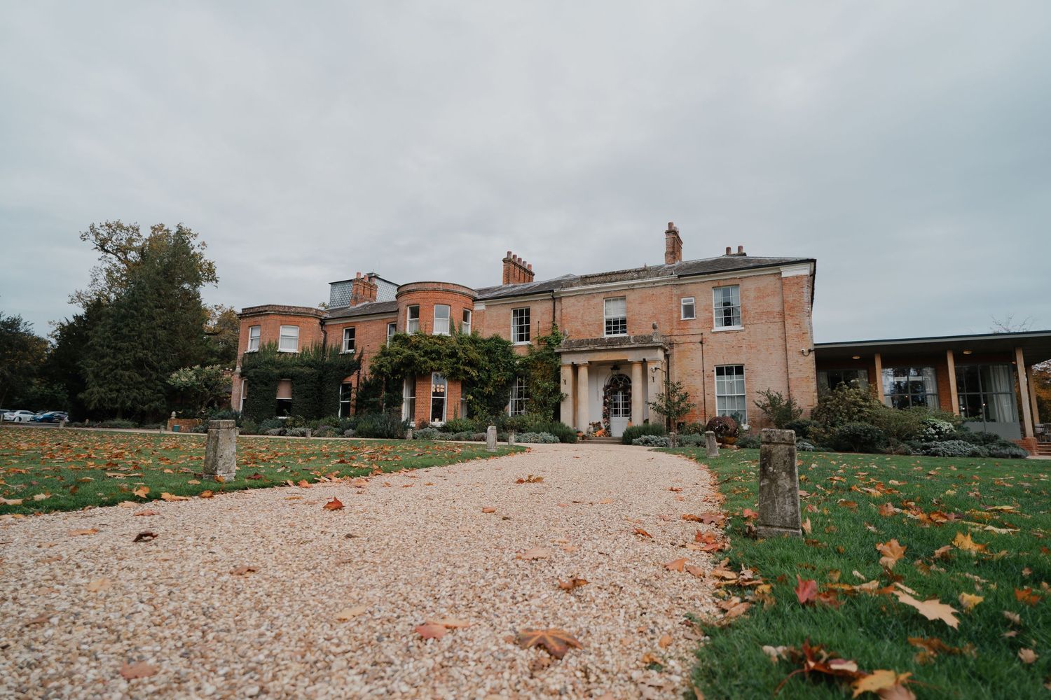 A grand Georgian country house with gravel driveway, ivy-clad walls, and autumn leaves on manicured grounds under overcast skies.