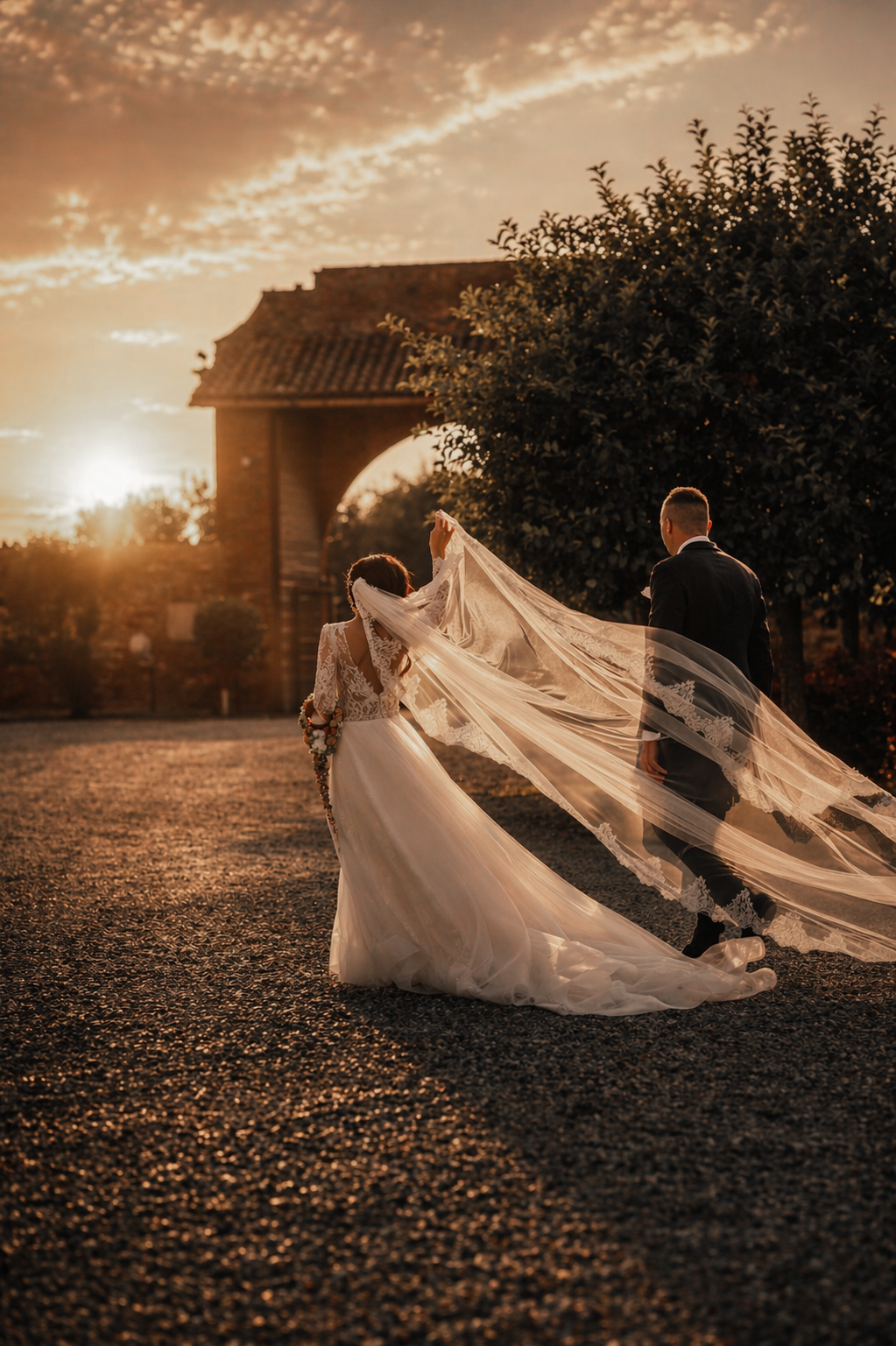 Bride in flowing gown with dramatic veil billowing in wind walking with groom at golden sunset.