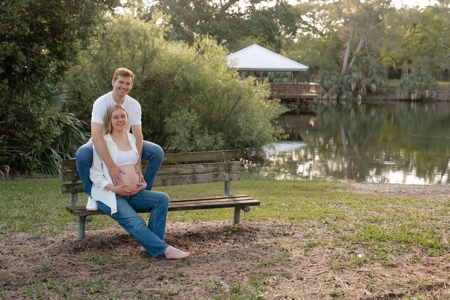 Couple posing on a park bench beside a serene pond surrounded by lush greenery during golden hour.