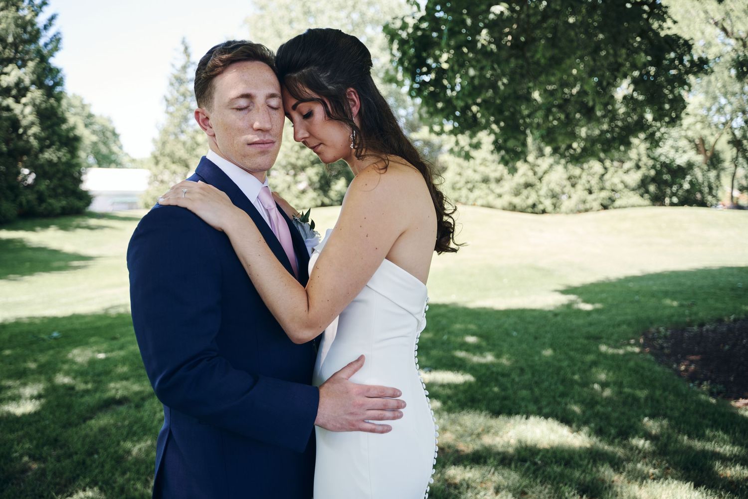 A bride and groom share a tender embrace outdoors on a sunny day, surrounded by lush green trees and grass.