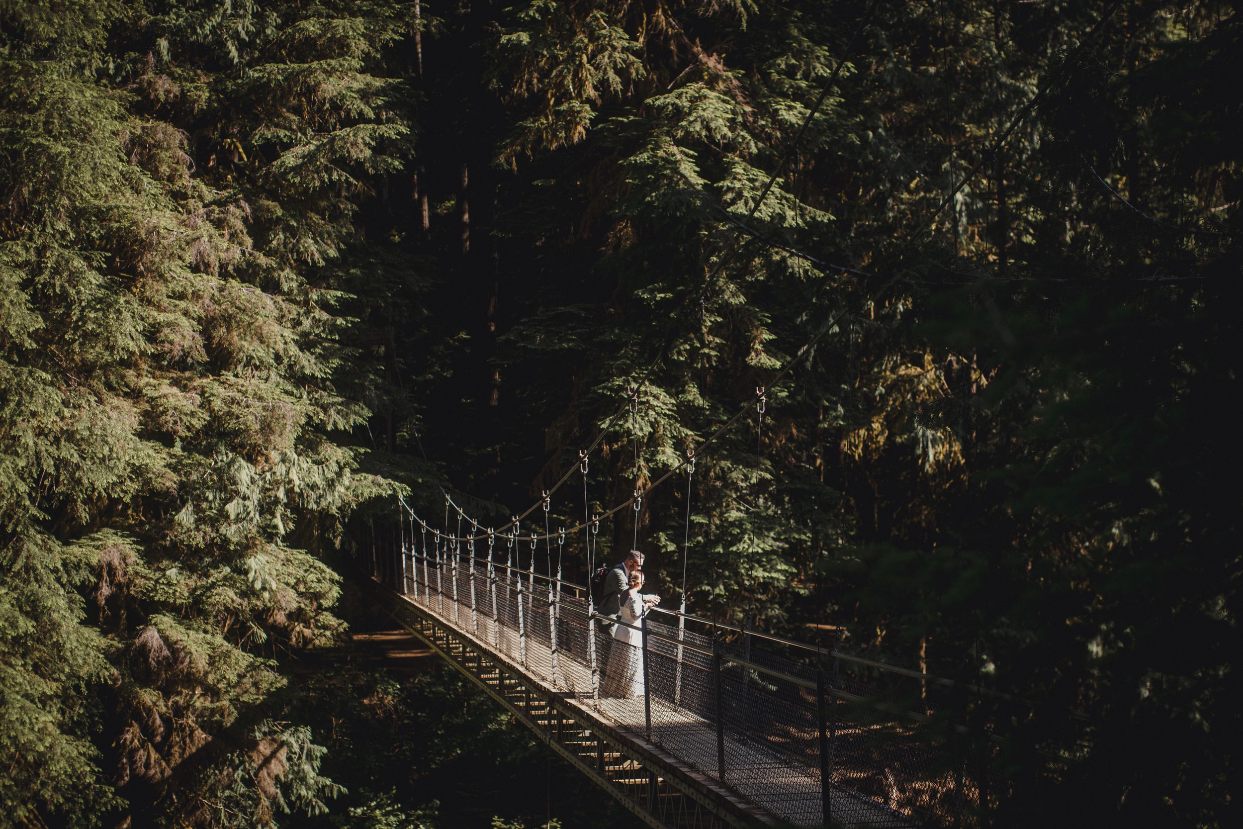 Bride and groom stand on a wooden suspension bridge surrounded by tall evergreen trees in a lush forest setting.