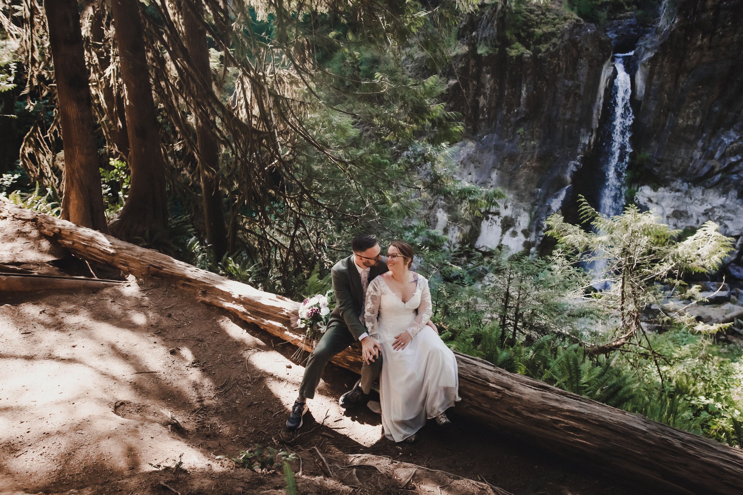 Bride and groom sitting on a fallen log in a forest with a waterfall in the background.