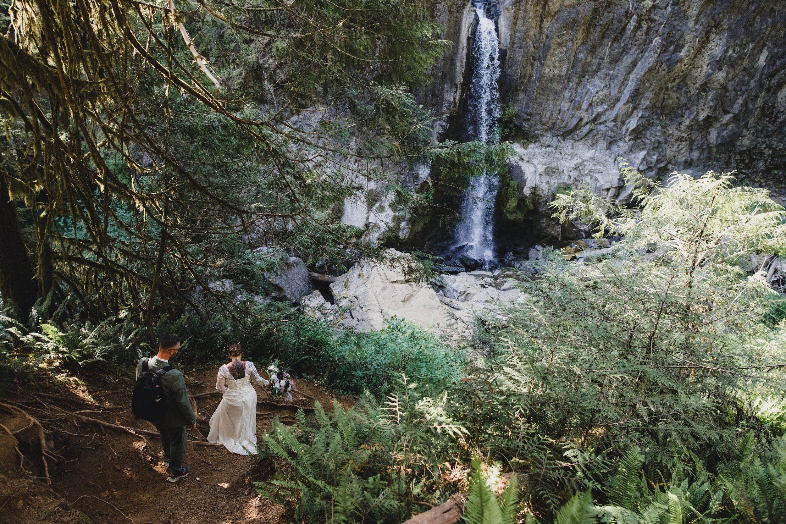 Wedding couple walks toward stunning waterfall surrounded by lush green forest.