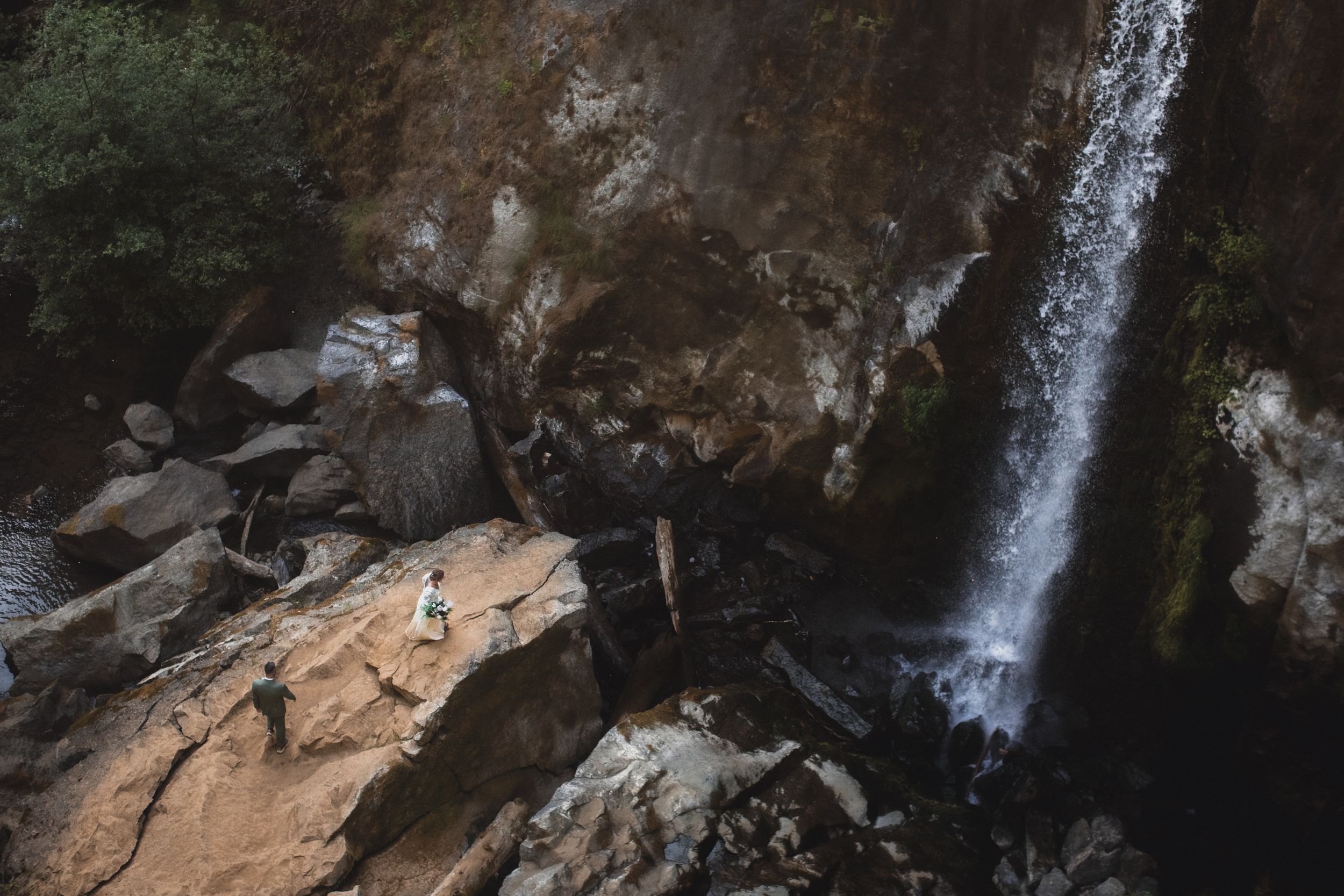 Couple stands on rocky terrain beside a dramatic waterfall cascading down a rugged cliff face.
