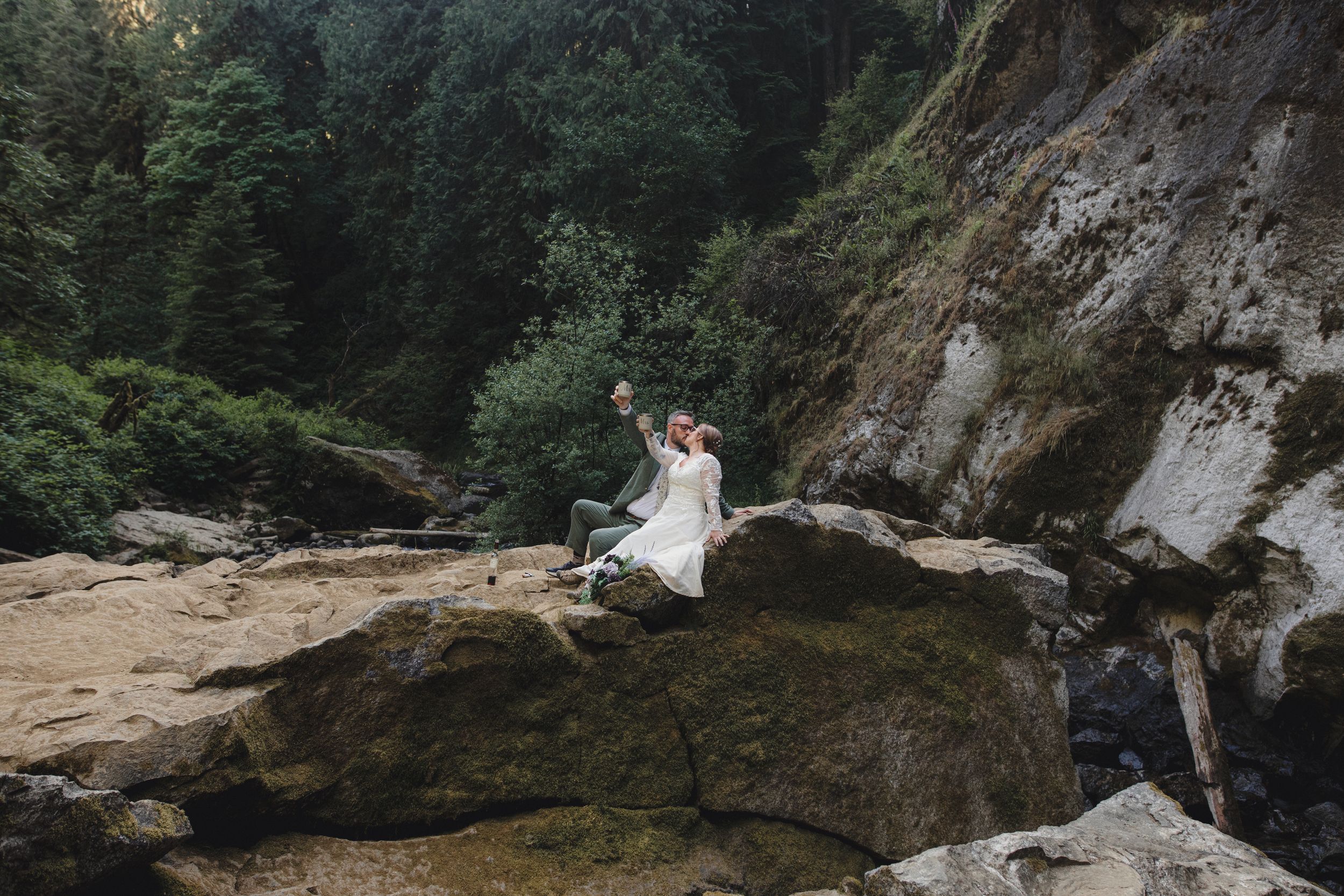 Couple in wedding attire sitting on mossy rocks surrounded by lush forest and rocky cliffs during outdoor elopement.