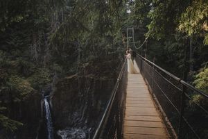 Couple walks on a wooden suspension bridge surrounded by lush forest with a waterfall below.