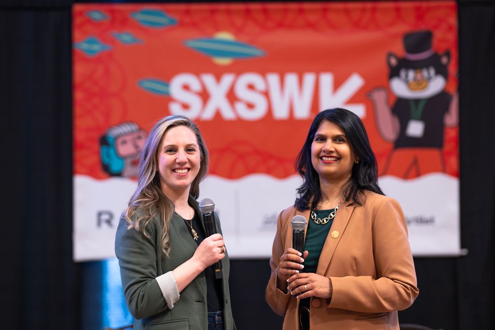 Two women smile and hold microphones on stage at SXSW event with colorful red branded backdrop.
