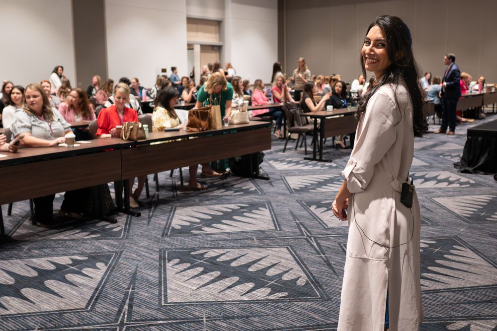A woman in a white outfit smiles at a conference room filled with seated attendees during a professional event.