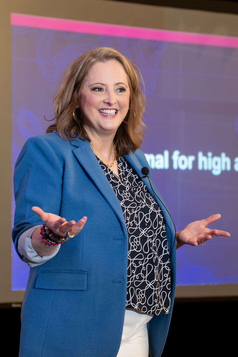 Woman in blue blazer smiling and gesturing while presenting at a professional conference event.