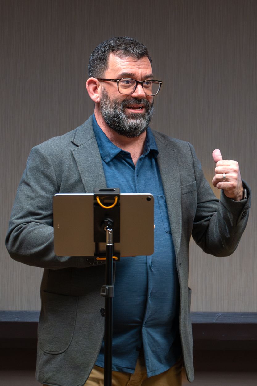 Bearded man in glasses and blazer giving a thumbs up while speaking at a podium with a tablet.