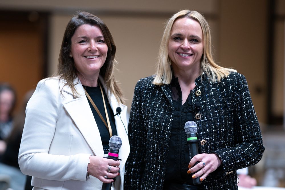 Two smiling women holding microphones on stage, one in white blazer and one in black patterned jacket.