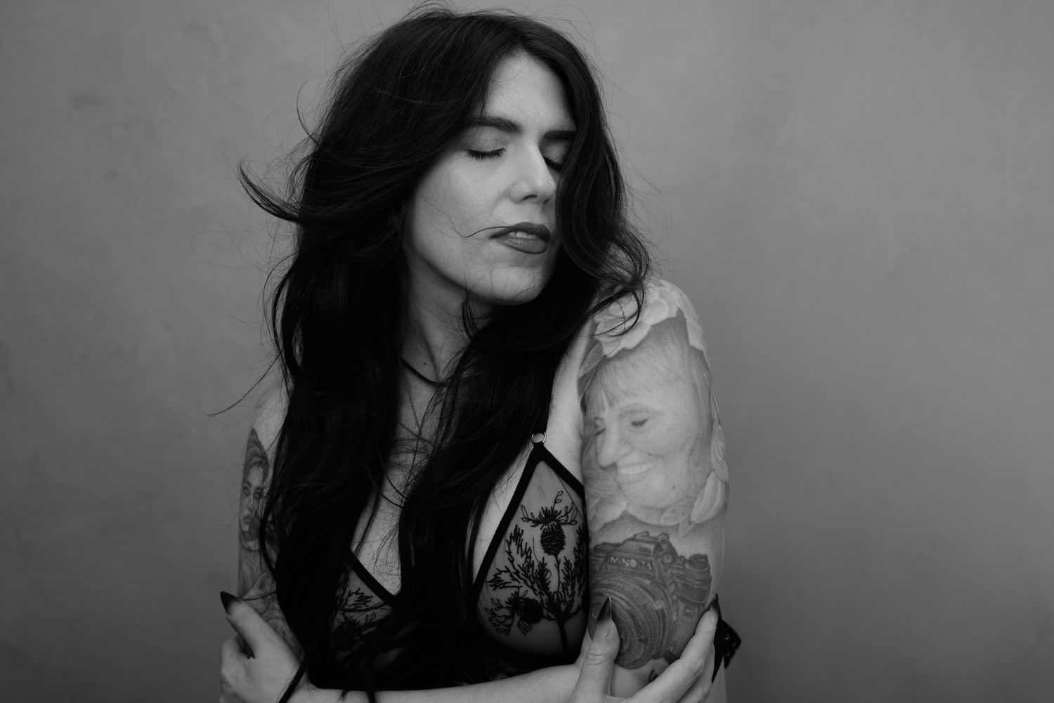 Black and white portrait of a tattooed woman with long dark hair, eyes closed, embracing herself gracefully.