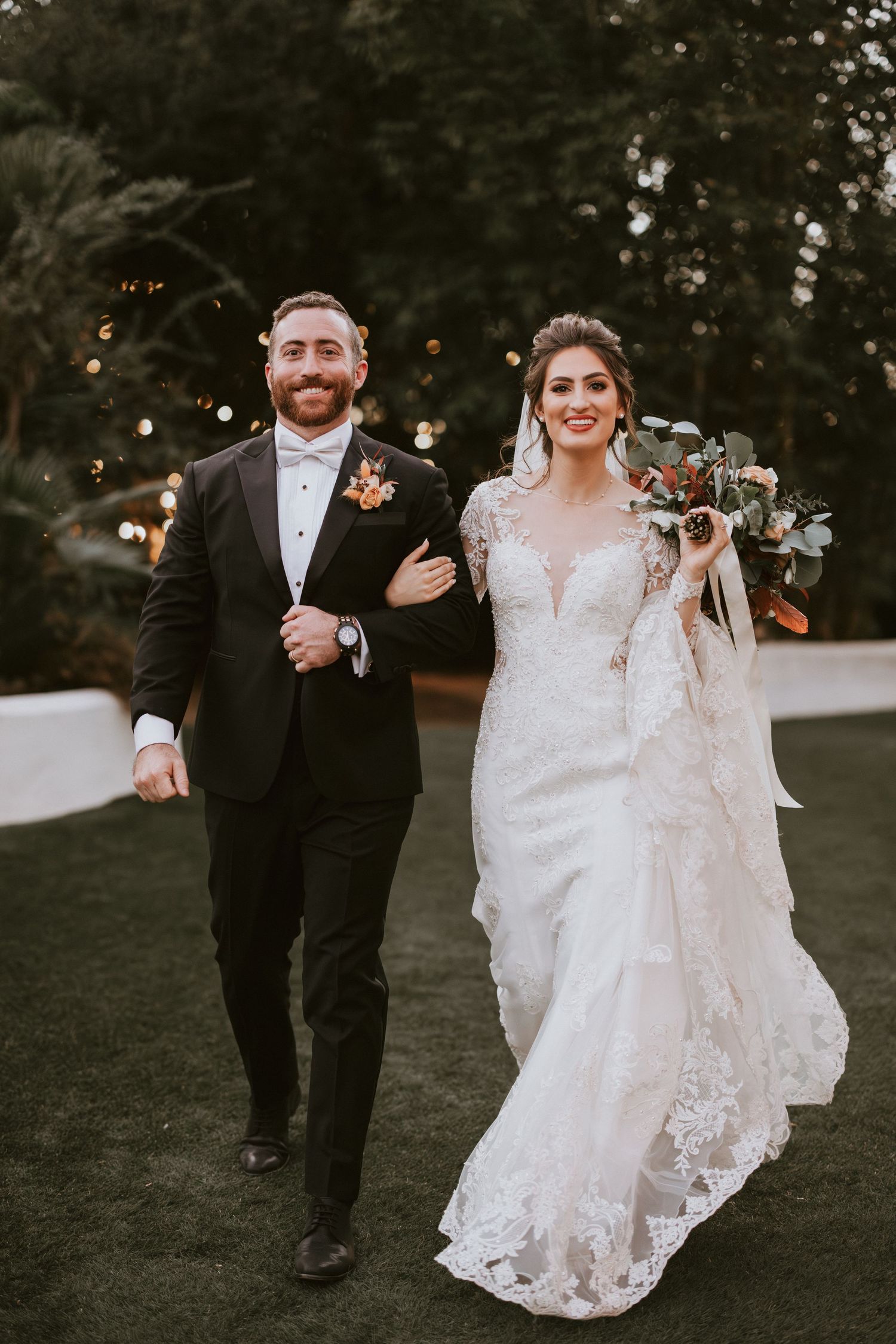 Bride in lace gown with train holds bouquet as groom in tuxedo kisses her cheek at outdoor wedding venue.