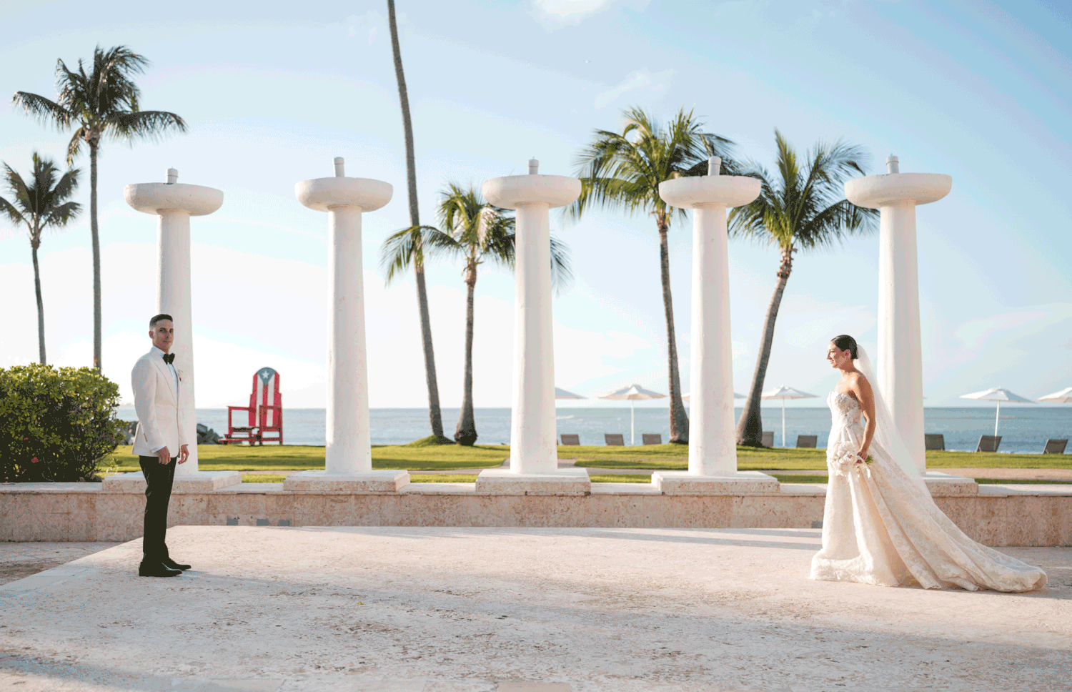 Bride and groom sharing a romantic moment under white columns with palm trees and ocean view in the background at Hyatt Regency Grand Reser
