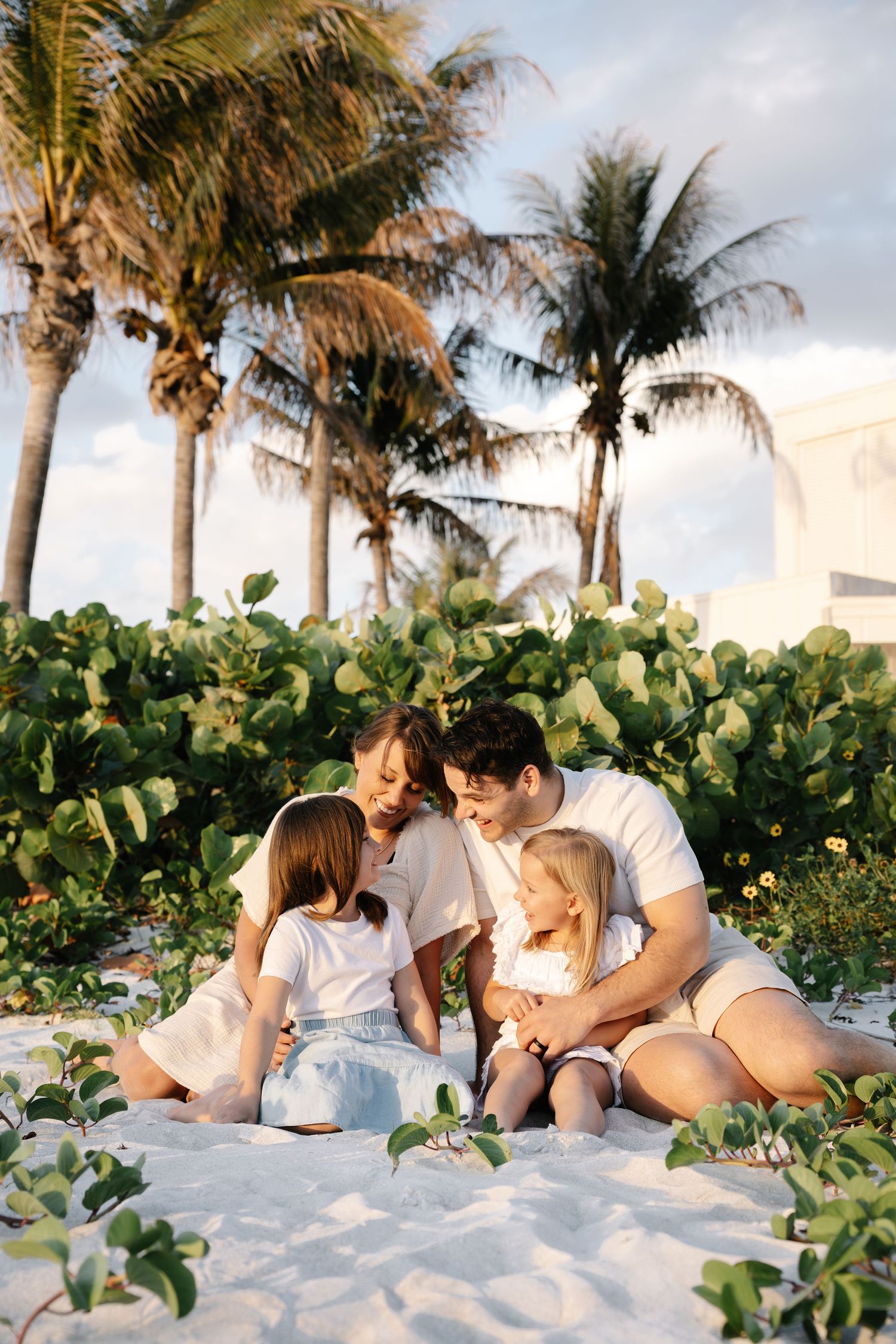 Family of four sitting together on white sand beach surrounded by tropical palms and lush greenery at golden hour.