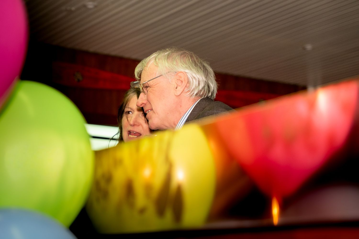 Elderly man and woman surrounded by colorful balloons at a festive indoor celebration event.