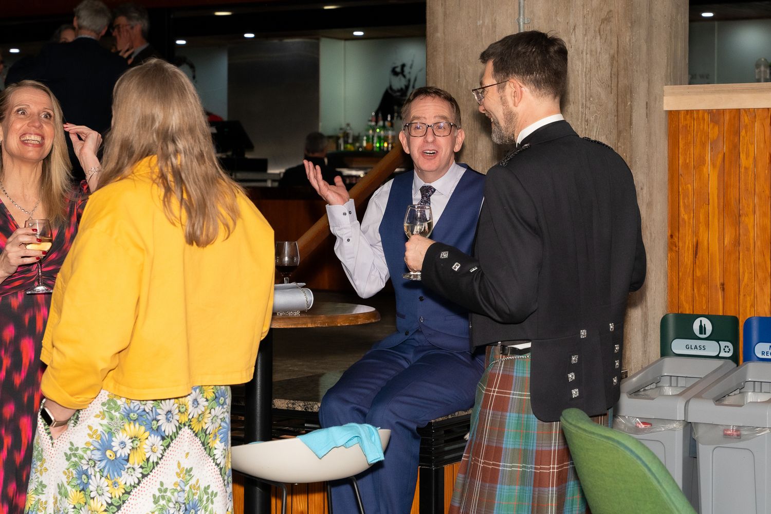 People socializing at a bar event, man in blue suit gesturing while talking to guests, another man in traditional Scottish kilt.