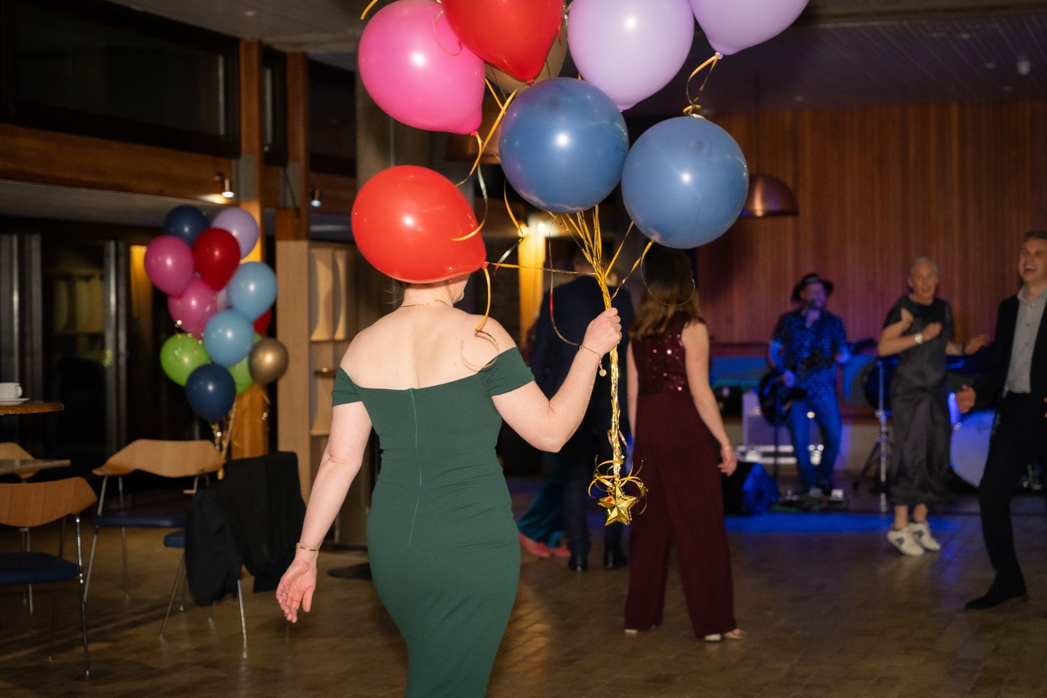 Woman in green dress carrying colorful balloons at an indoor party celebration with band performing in background.