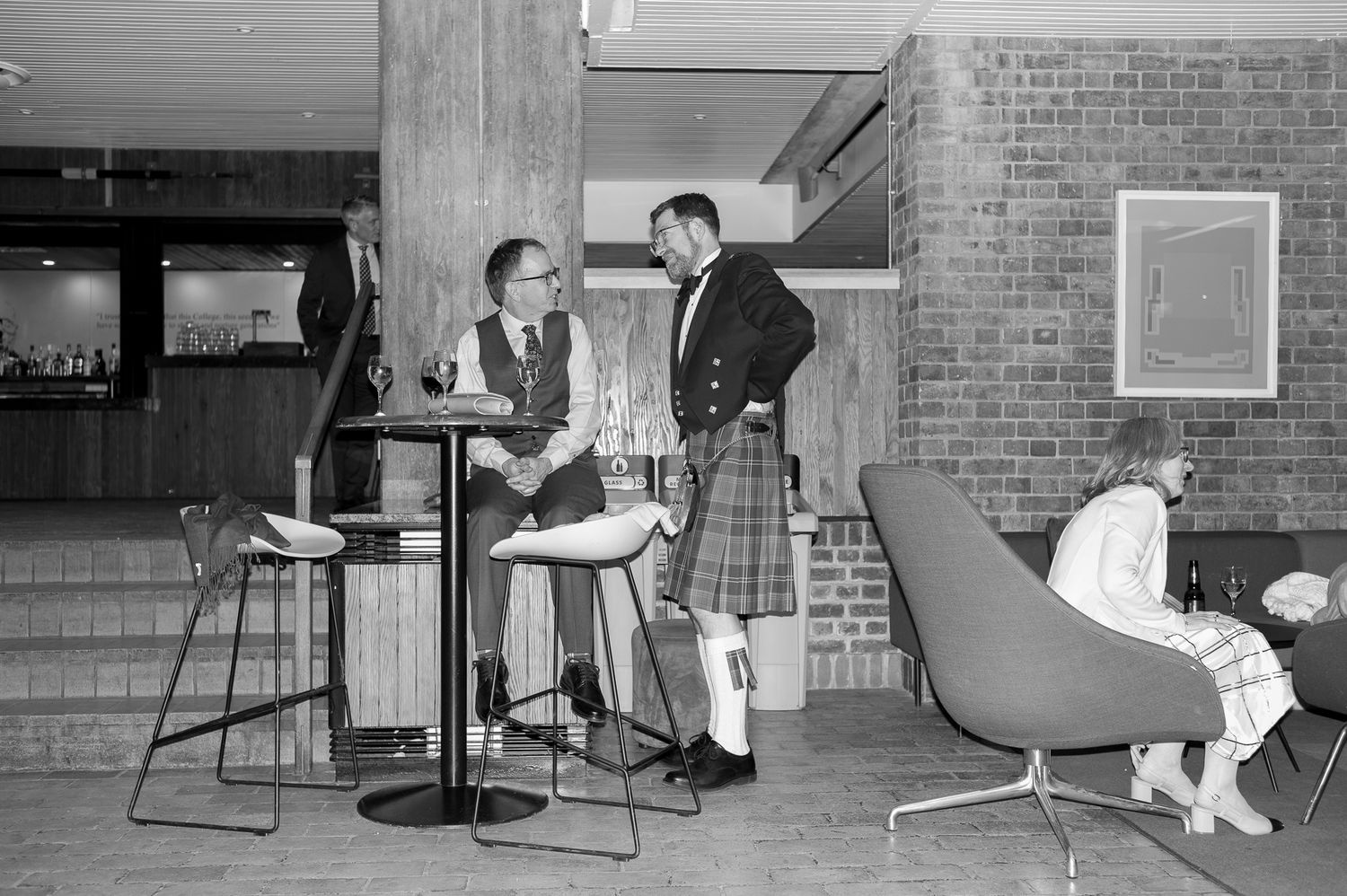 Black and white photo of two men in kilts conversing at a tall table in a mid-century modern lounge setting.