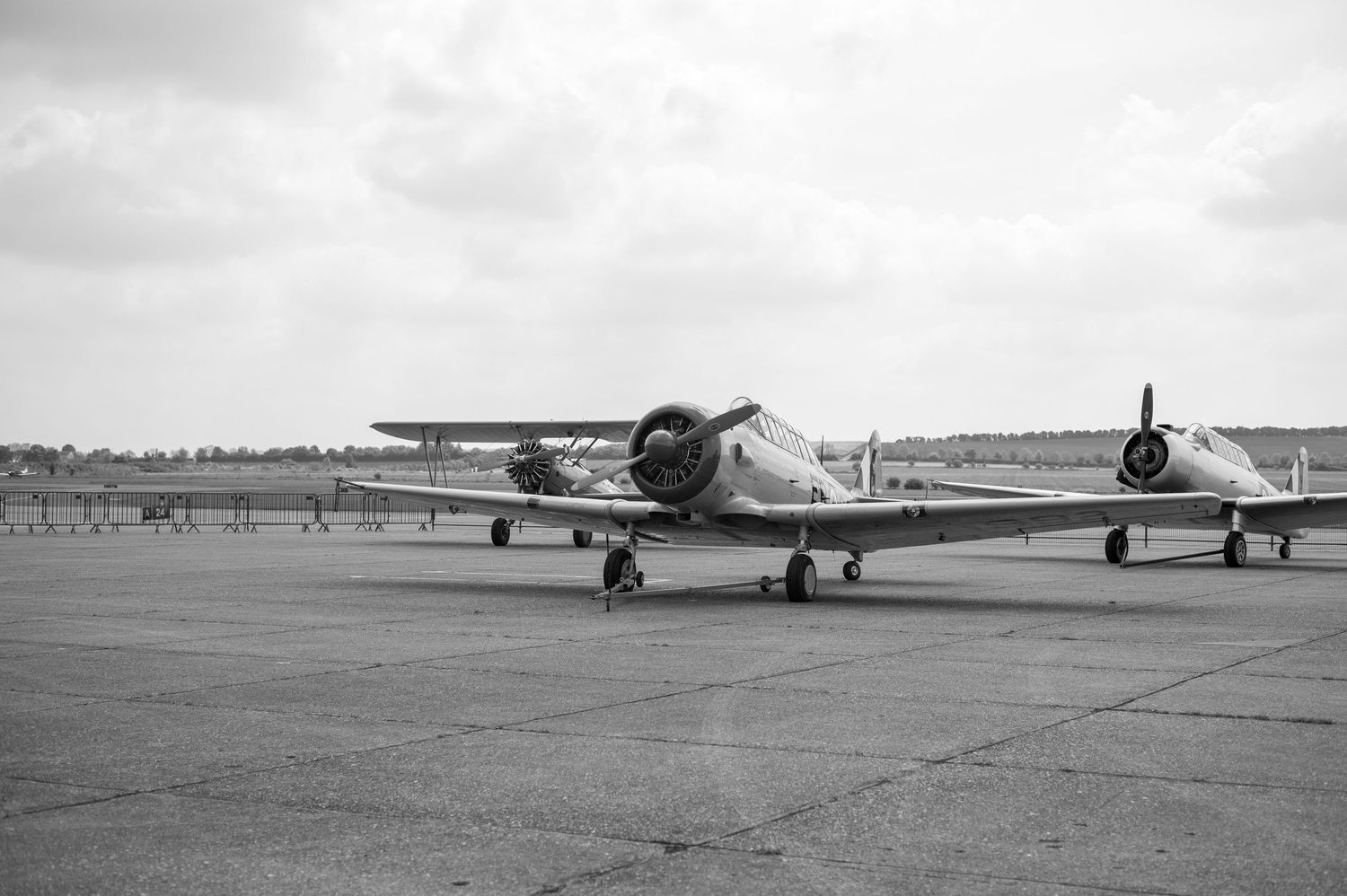 Black and white photo of vintage propeller aircraft parked on an airfield tarmac under a cloudy sky.