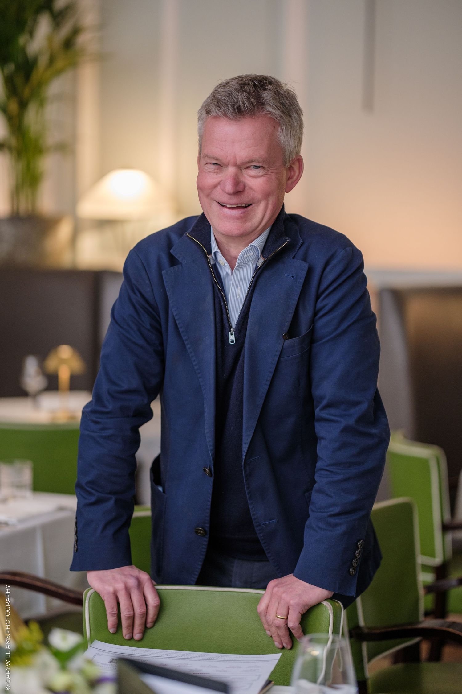 Soren Jessen in navy blazer leans on green chair in elegant restaurant setting.