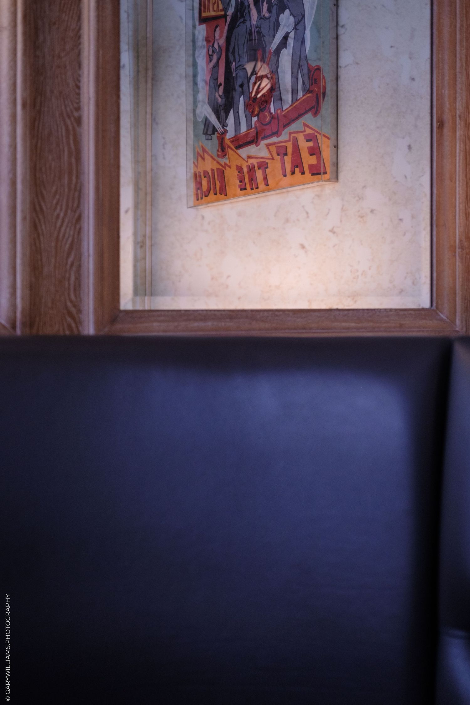 Vintage framed poster reading 'Eat The Rich' displayed above a dark leather booth seat in a restaurant.