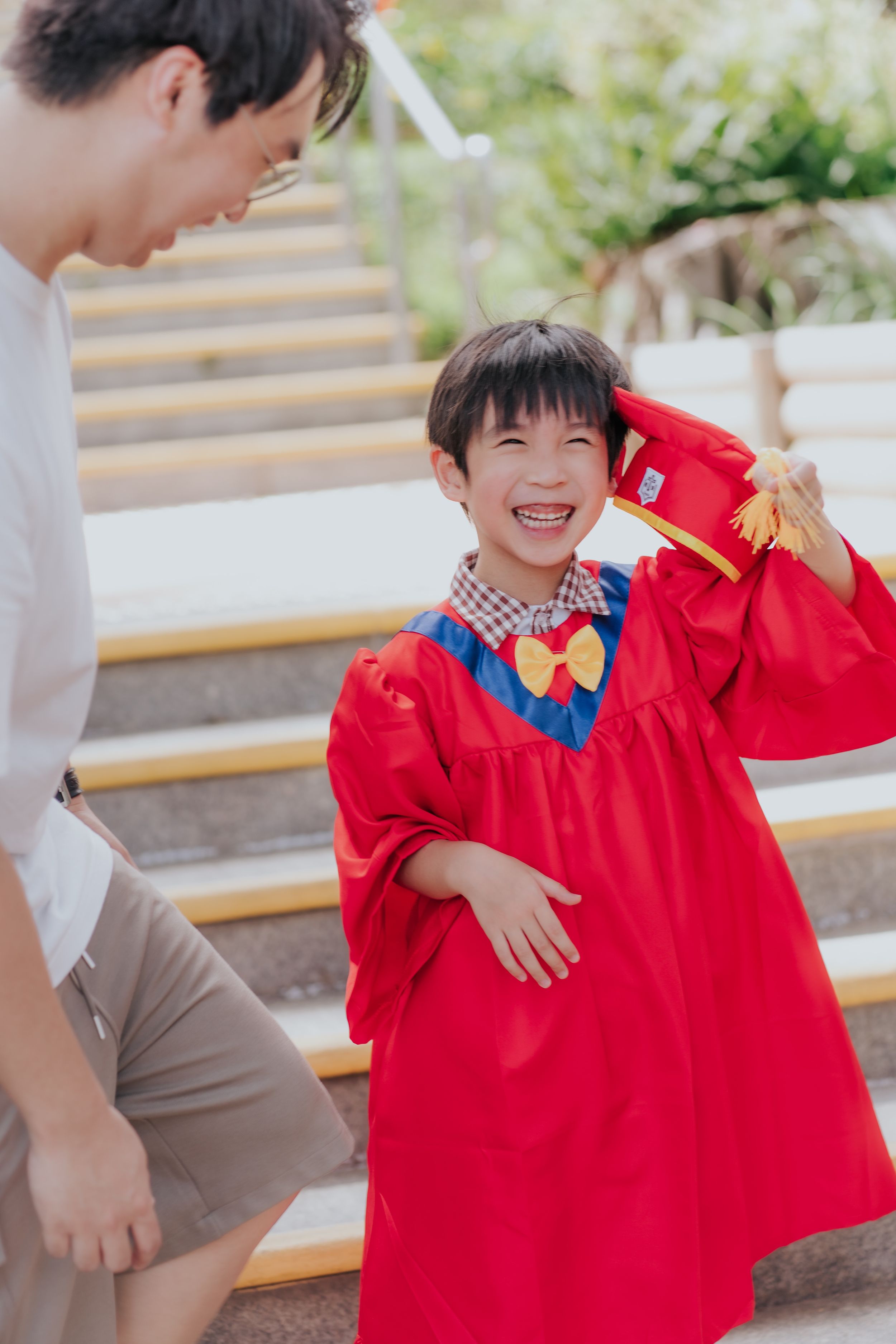 Happy child in red graduation gown and cap smiling with parent on steps during kindergarten ceremony.一位快樂的兒童身穿紅色畢業禮服，戴著畢業帽，在幼兒園畢業典禮上與父母在微笑合影