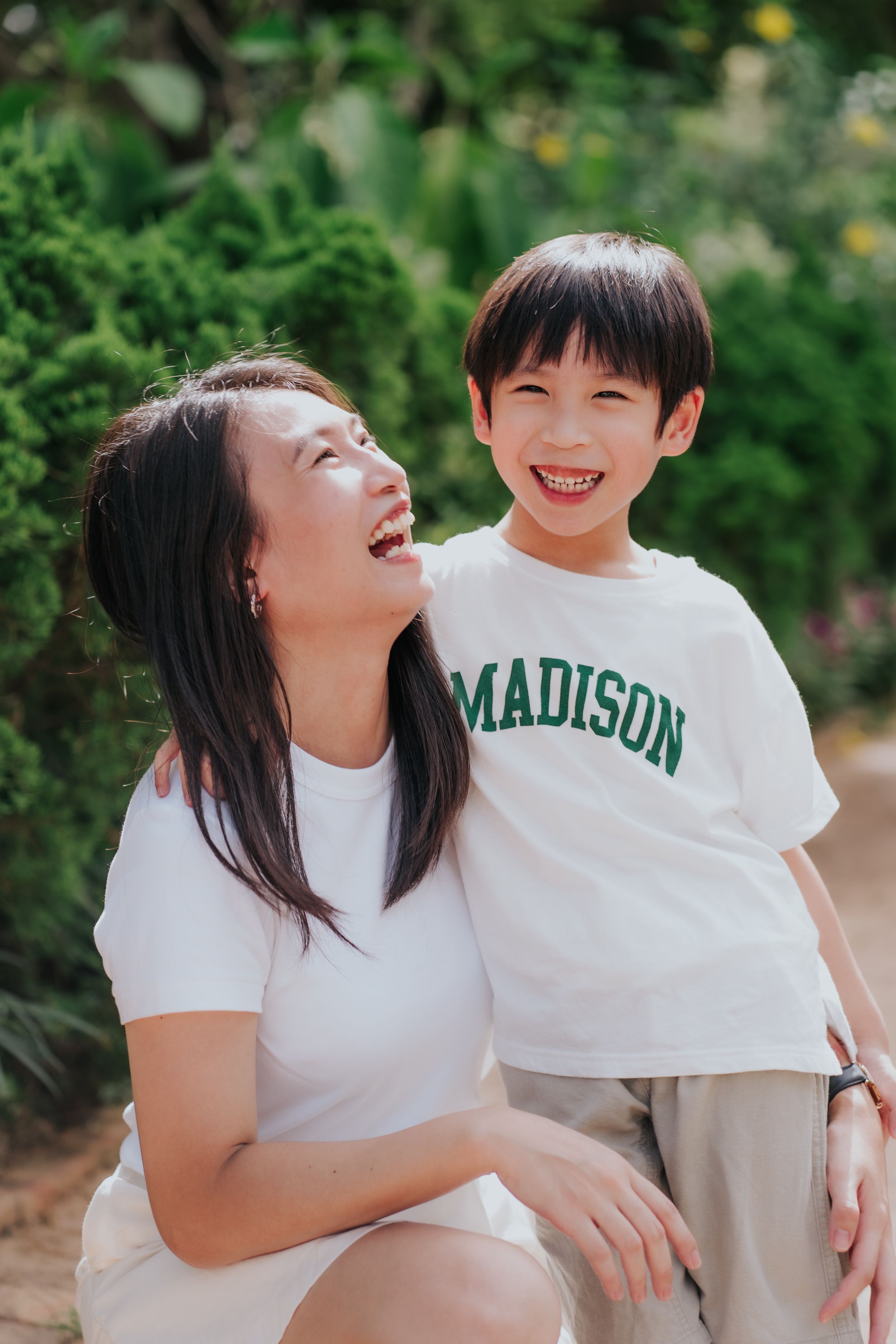A smiling Asian mother and son in white t-shirts share a joyful moment outdoors surrounded by green foliage.
一位笑容燦爛的亞裔母親和她的兒子穿著白色T卹，享受著快樂的時光