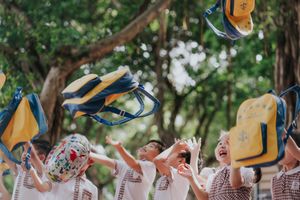 Children joyfully tossing blue and yellow backpacks into the air outdoors under green trees.
孩子們在綠樹成蔭的戶外，興高采烈地將藍色和黃色的書包拋向空中。