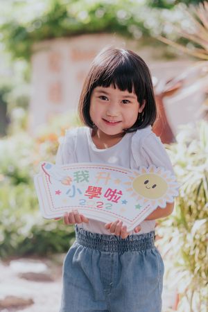 Young Asian girl smiling and holding a colorful 2022 elementary school sign outdoors in a sunny garden.
一位年輕的亞洲女孩面帶微笑，在陽光明媚的戶外花園裡舉著一塊色彩鮮豔標誌牌