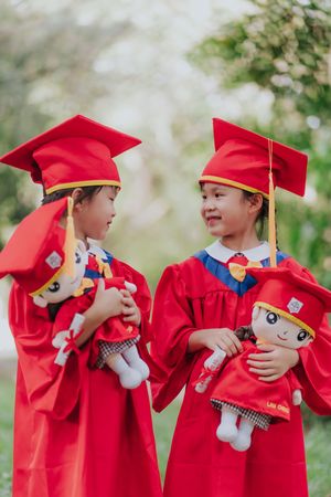 Two young children in red graduation gowns and caps hold matching dolls outdoors.兩個穿著紅色畢業禮服、戴著畢業帽的小孩在戶外抱著同款娃娃。
