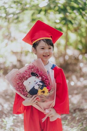 Smiling young girl in red graduation cap and gown holding a pink flower bouquet outdoors.
一位面帶笑容的年輕女孩身穿紅色畢業禮服，頭戴畢業帽，手捧粉色花束，在戶外。