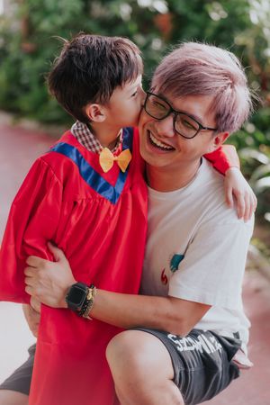 A young boy in red graduation gown kisses a laughing adult with pink hair and glasses outdoors.
一個身穿紅色畢業禮服的小男孩在戶外親吻一個戴著眼鏡、留著粉紅色頭髮的成年人，成年人正笑著