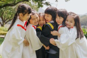 Group of young children in graduation gowns hugging and celebrating outdoors on a sunny day.一群穿著畢業禮服的小朋友在陽光明媚的日子裡戶外擁抱慶祝。