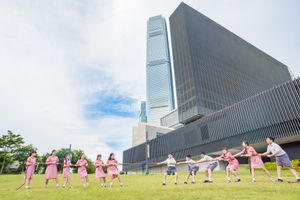 Children in pink uniforms play on green grass in front of modern skyscrapers on a sunny day.
在陽光明媚的日子裡，一群穿著粉紅色制服的孩子在現代摩天大樓前的綠草地上玩耍。
