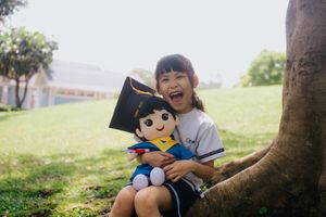 A happy girl hugs a graduation-themed plush toy while sitting by a tree in a sunny park.一位快樂的女孩坐在陽光明媚的公園裡的一棵樹旁，抱著一個畢業主題的毛絨玩具。