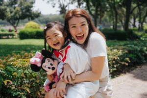 A joyful mother and daughter laugh together outdoors, the girl holding a Minnie Mouse plush toy.一對快樂的母女在戶外一起開懷大笑，女孩手裡拿著米妮老鼠毛絨玩具。