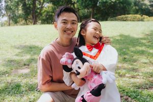 A smiling young man and laughing girl pose outdoors holding a Minnie Mouse plush toy in a green park.一位面帶笑容的年輕男子和一位開懷大笑的女孩在綠樹成蔭的公園裡，手捧米妮老鼠毛絨