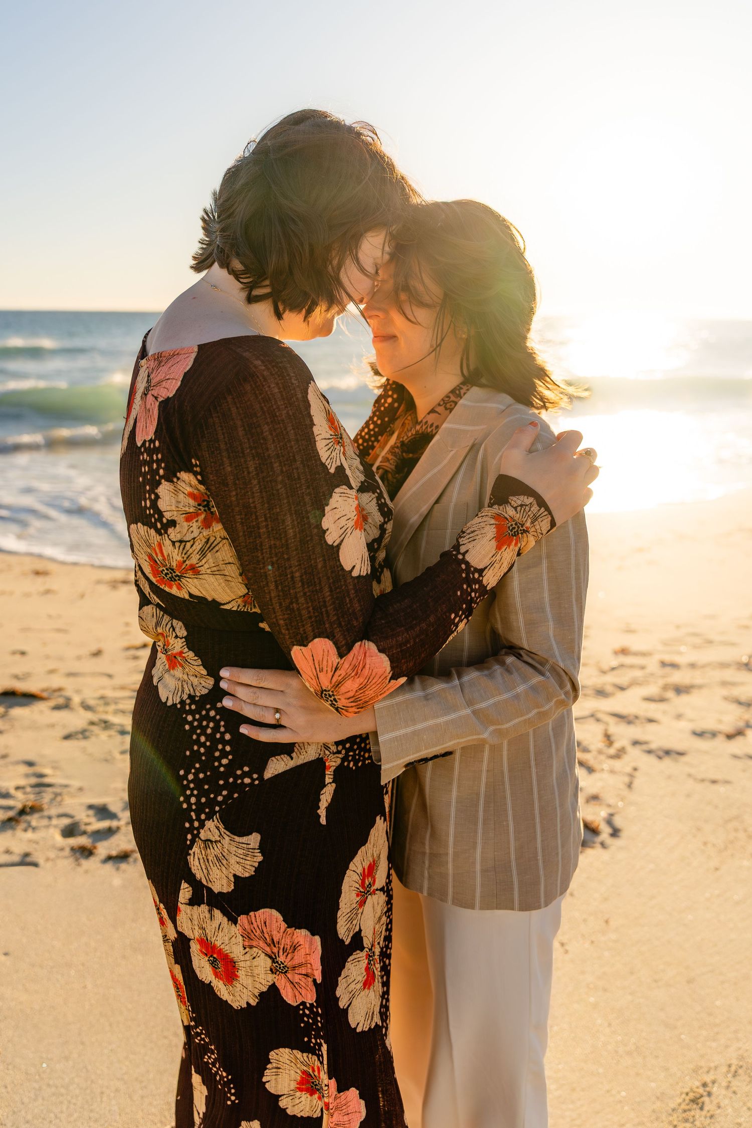 Couple embracing on a sunny beach at golden hour, wearing floral dress and striped blazer, with ocean waves in the background.