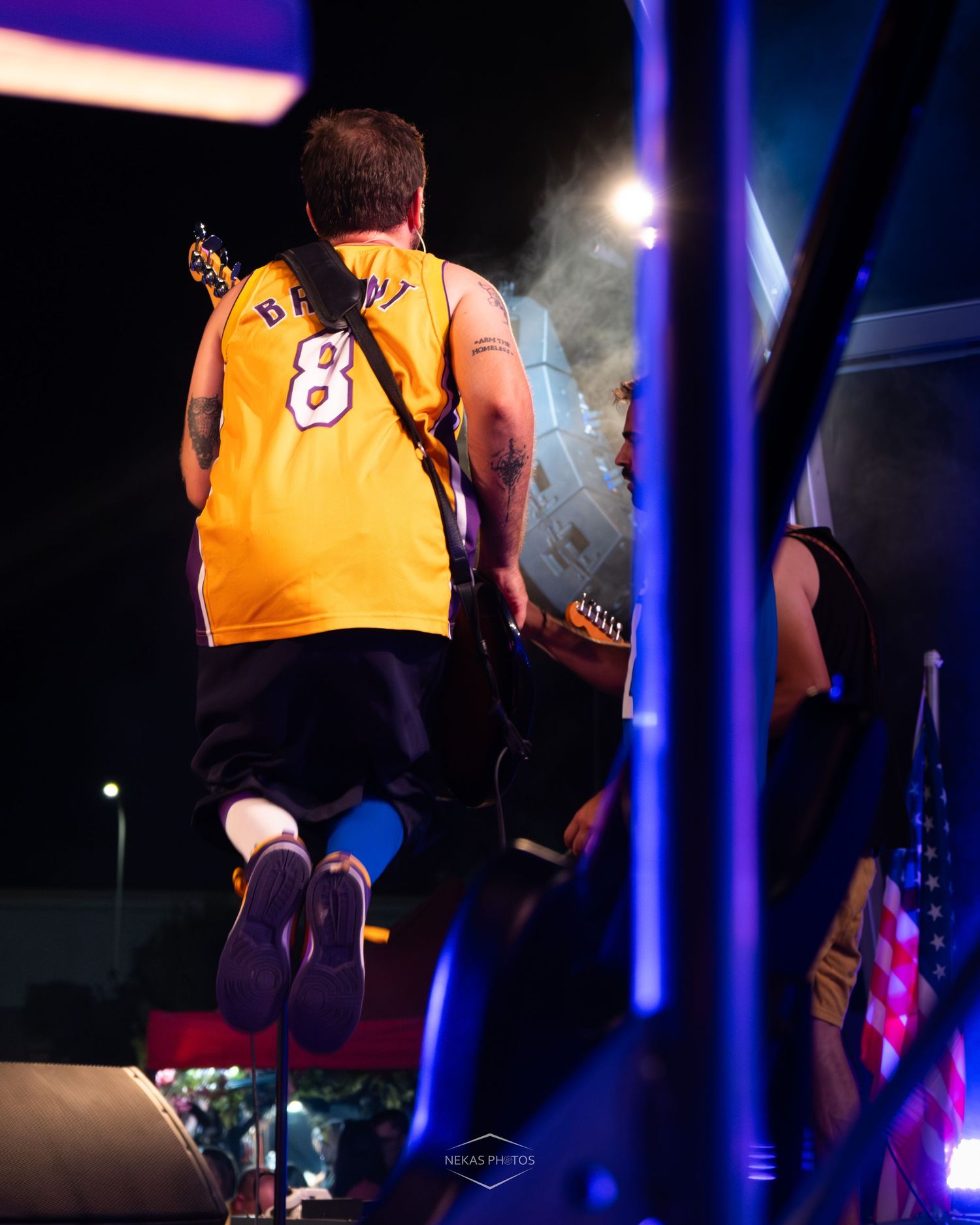 Guitarist in a yellow Lakers #8 Bryant jersey jumps on stage during a live outdoor concert at night.