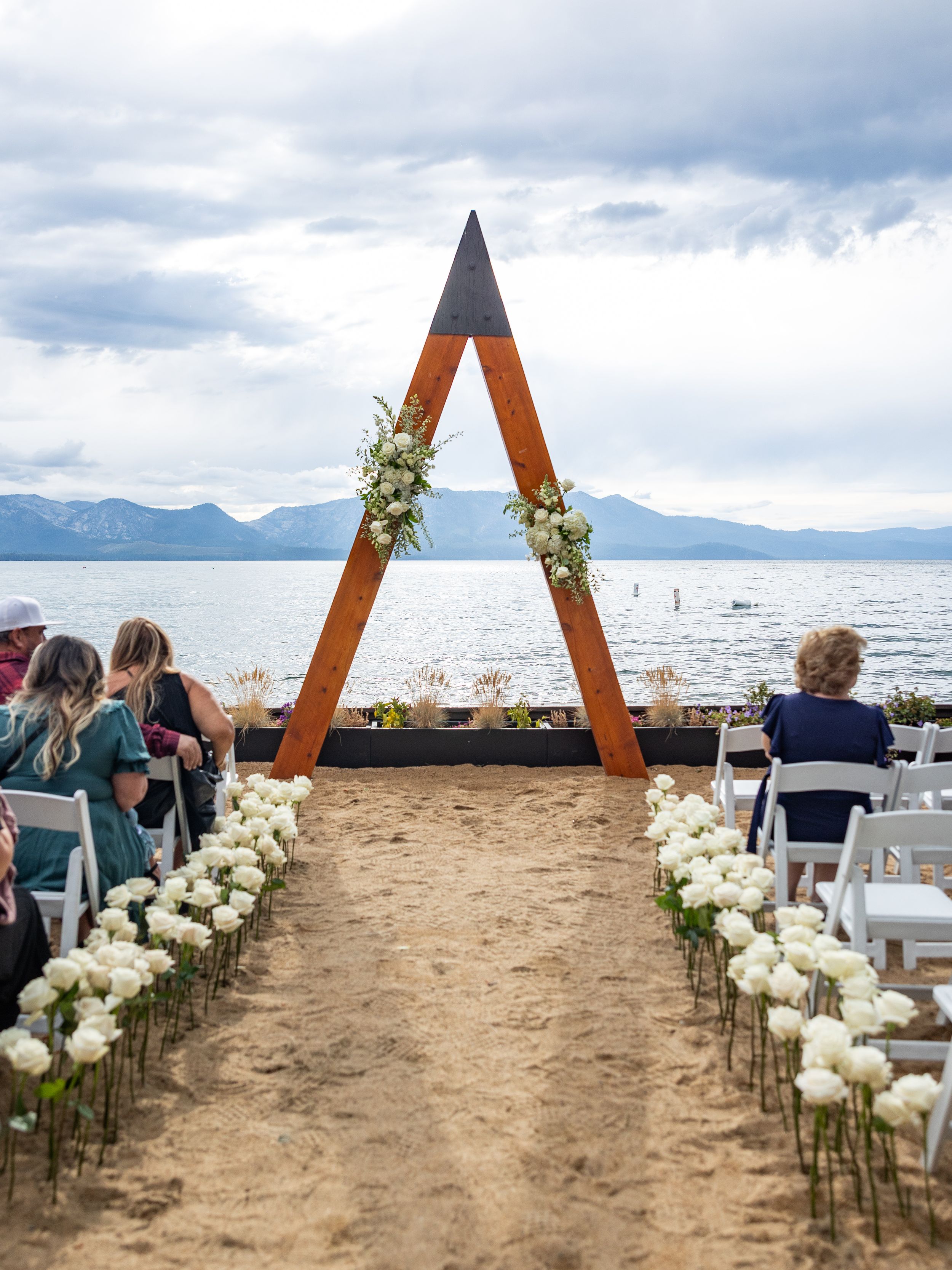 Outdoor beach wedding ceremony with white roses lining the aisle, triangular wooden arch, and Lake Tahoe mountain views.