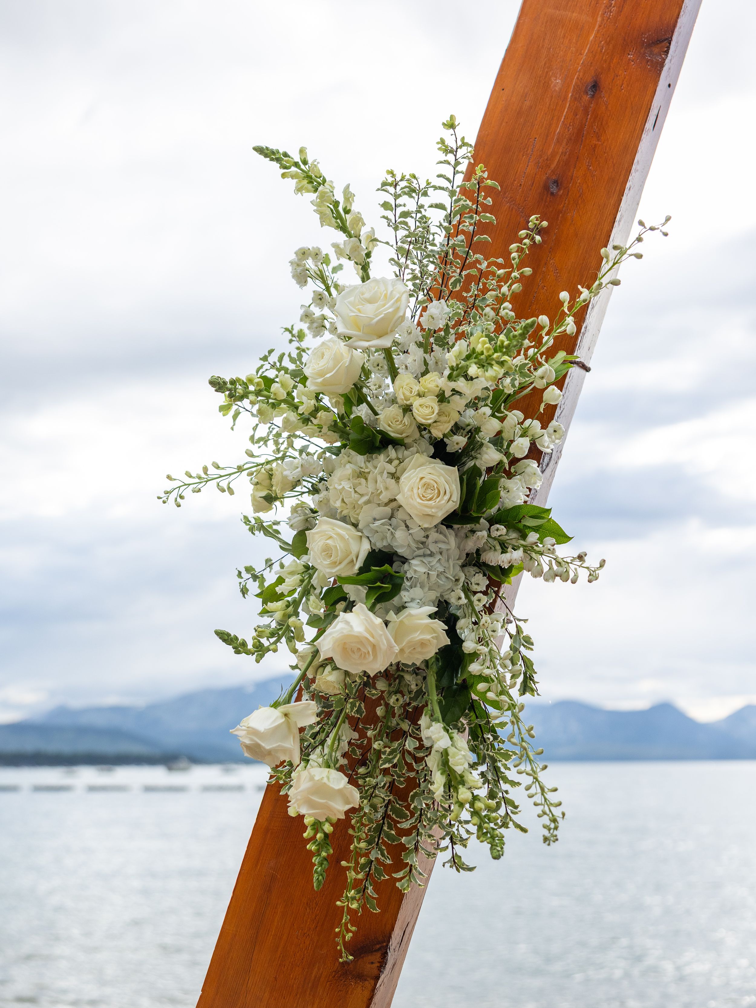 White roses and greenery floral arrangement on a wooden arch with a lake and mountains in the background.