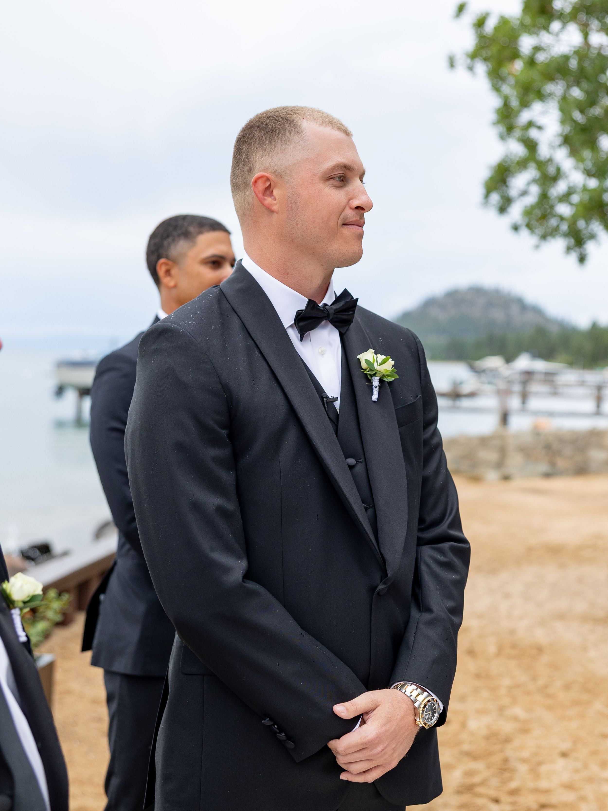Groom in black tuxedo with boutonniere stands at outdoor lakeside wedding ceremony, looking emotional.