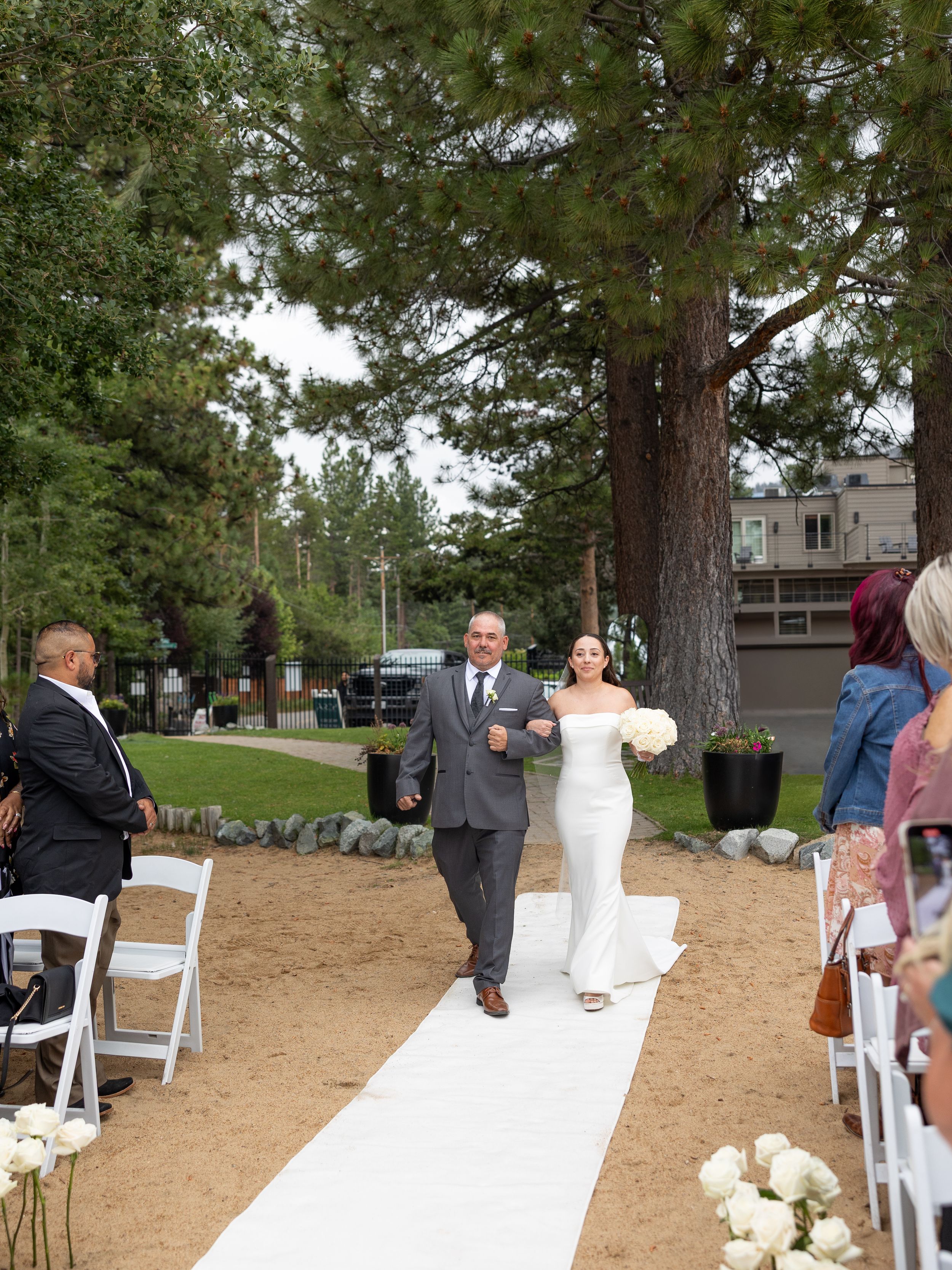 Bride in white gown walks down sandy aisle with escort at outdoor wedding ceremony surrounded by pine trees.