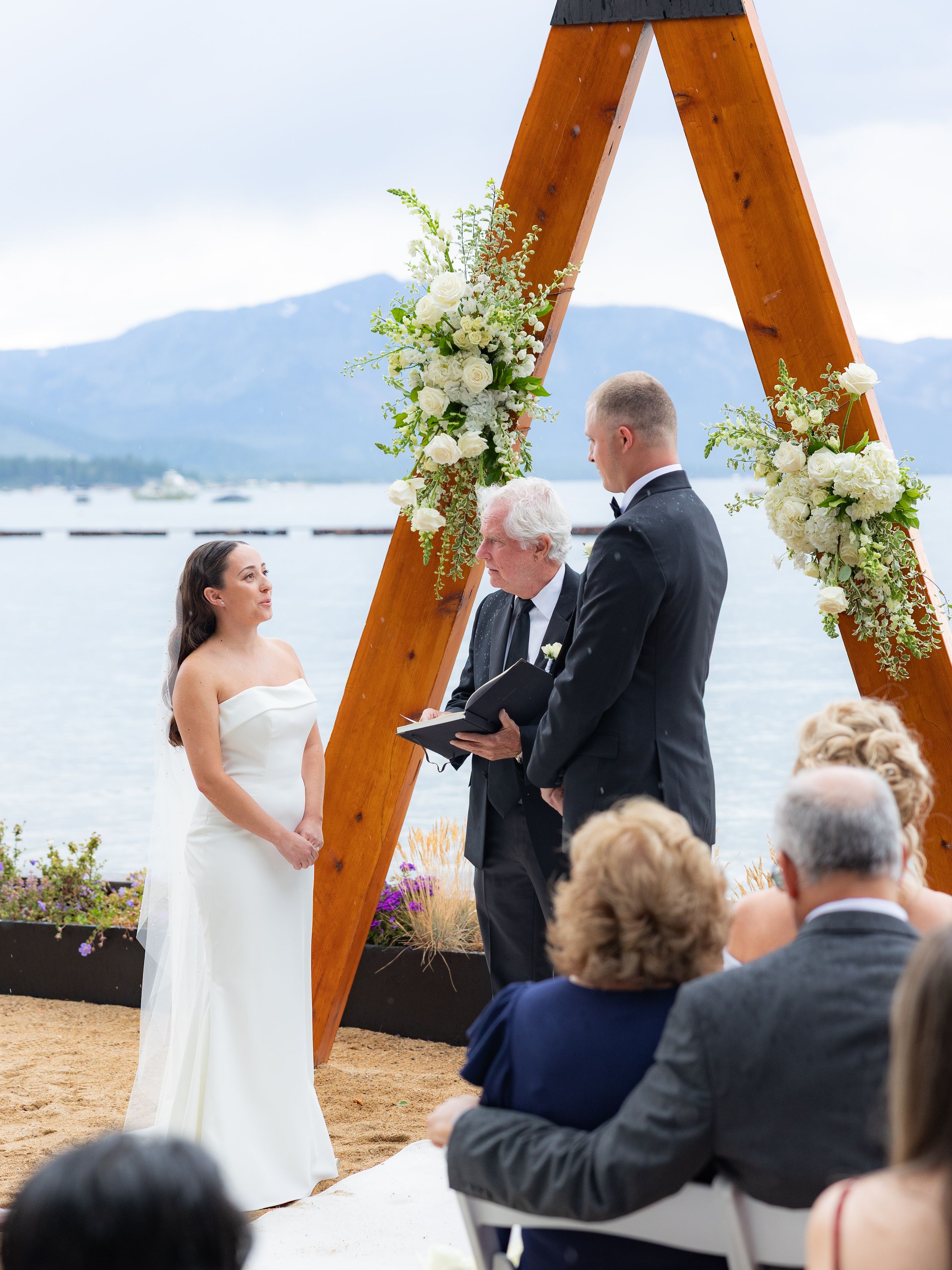 Bride and groom exchange vows under a wooden arch with white flowers by Lake Tahoe.