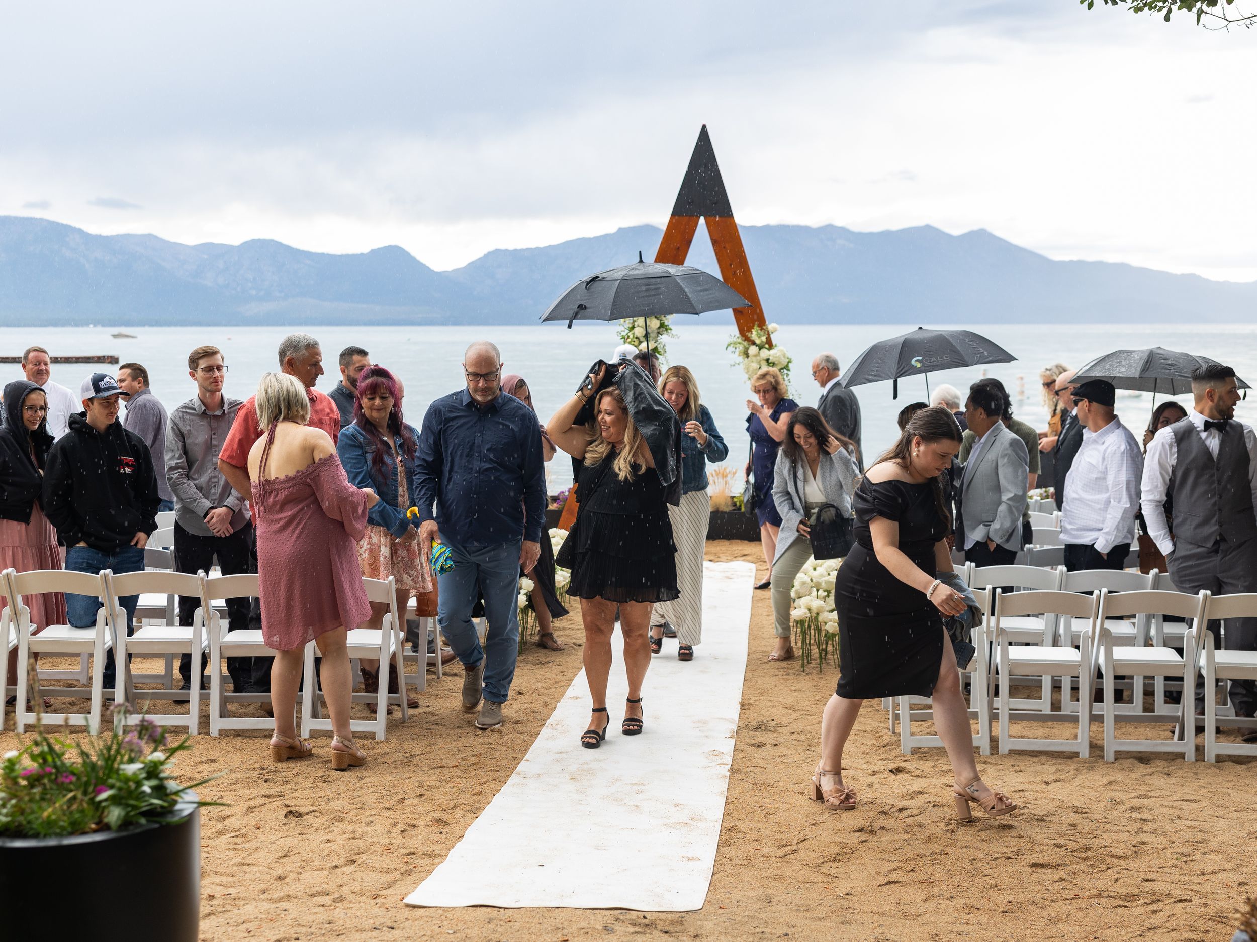 Outdoor beach wedding ceremony at Lake Tahoe with guests seated in white chairs under cloudy skies.