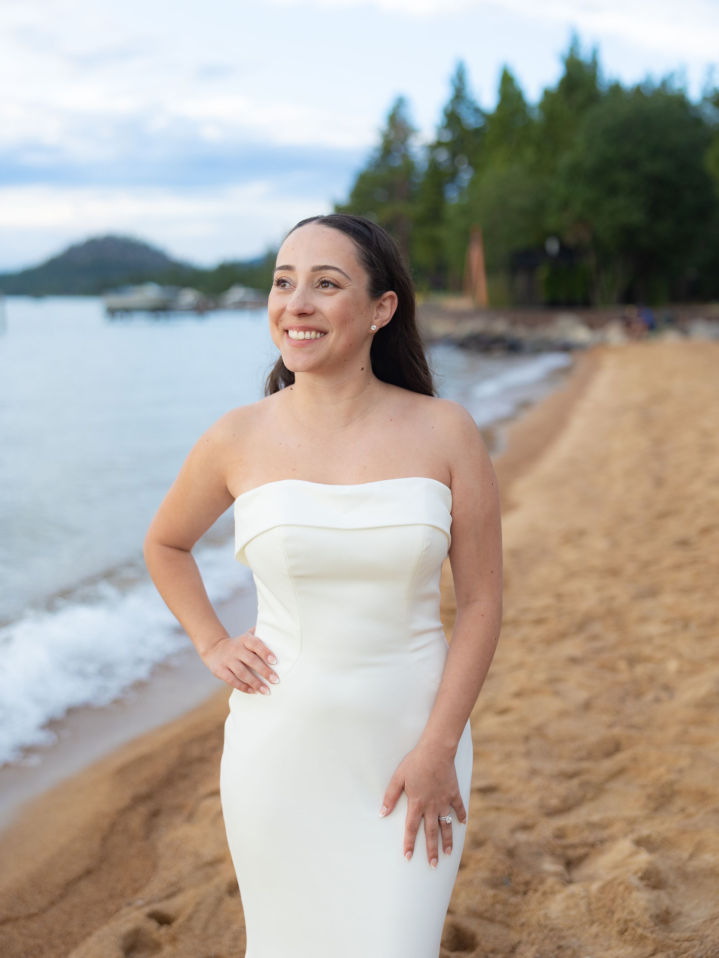 Woman in white strapless dress smiling on sandy beach with ocean and trees in background.