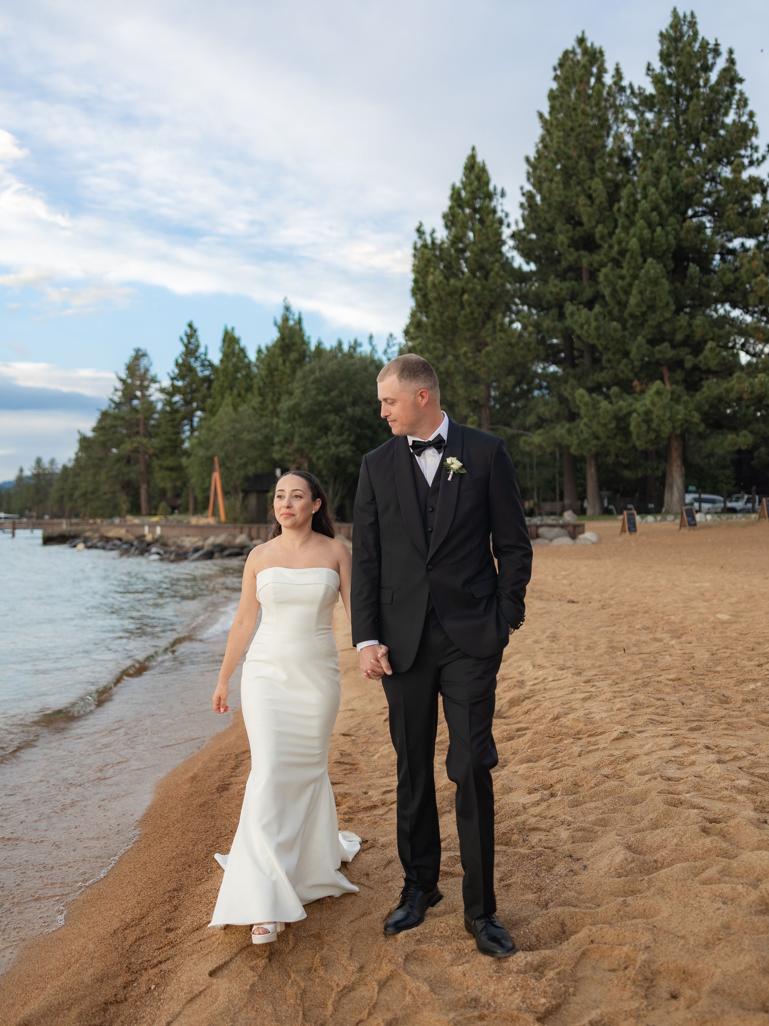 Bride and groom walking hand in hand on a sandy beach at Lake Tahoe in wedding attire.