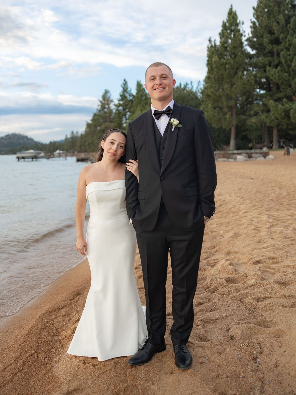 Newlywed couple posing together on a sandy Lake Tahoe beach, bride in white gown and groom in tuxedo.